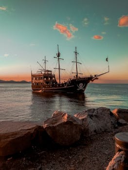 A pirate ship sails during sunset, casting a dramatic shadow on calm ocean waters against a colorful sky.